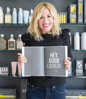 Alli Webb in a Drybar holding an open book that reads, "Hey, good lookin." 