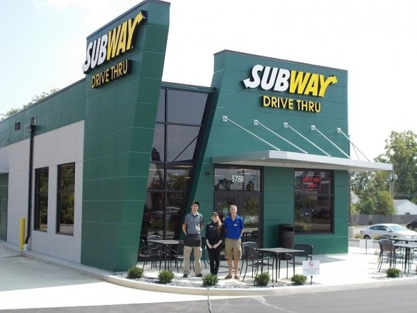 Image of a Subway restaurant with a dark green exterior and a drive thru, with 3 restaurant employees standing outside