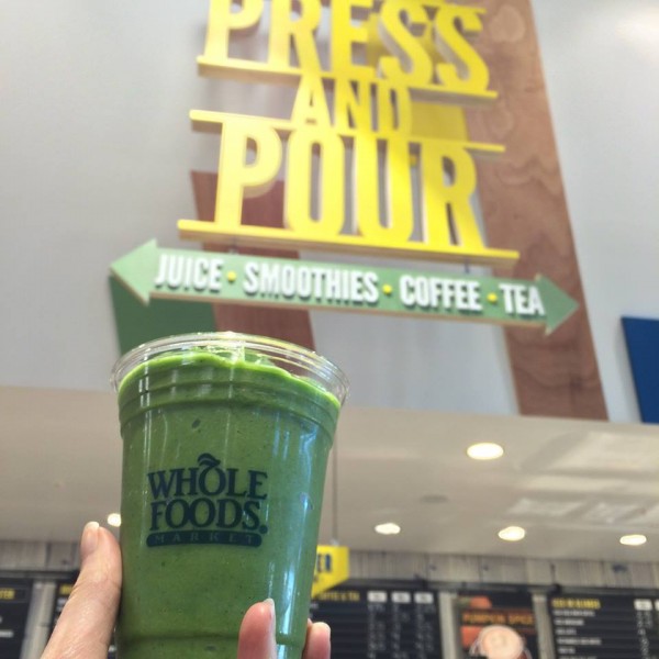 A green smoothie in a Whole Foods cup begin held up in front of a whole foods press and pour section