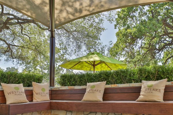 Outdoor seating area at Westlake Plaza under a canopy with four pillows on wooden benches in front of shrubs and trees