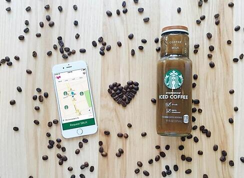 Overhead view on an iPhone, a bottle of cold coffee from starbucks, and coffee beans shaped like a heart, all on a wooden table. 