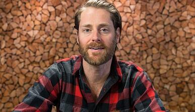 Bearded man in flannel shirt in front of a wall of chopped wood