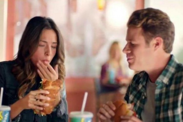 screenshot of a tv commercial where man and woman are eating subway sandwiches inside of a subway restaurant