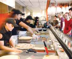 Employees adding toppings to pizzas for a line of customers. 