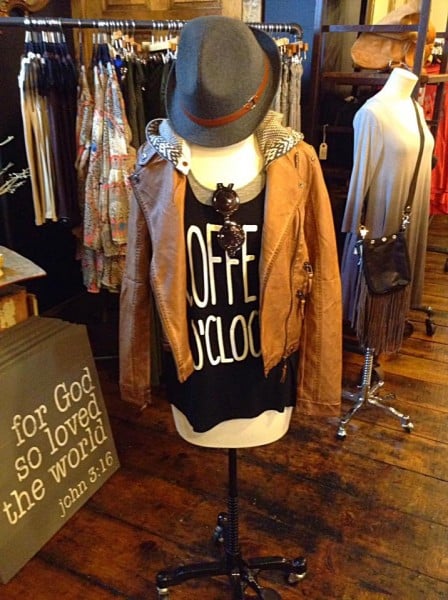 a mannequin displaying a black shirt that says "coffee o'clock", a tan leather jacket, and a gray fedora hat