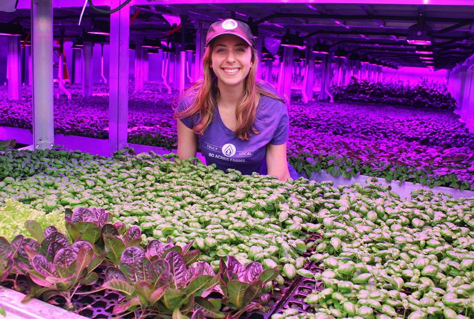 80 Acres Farms employee posing in a greenhouse lit by purple lights. 