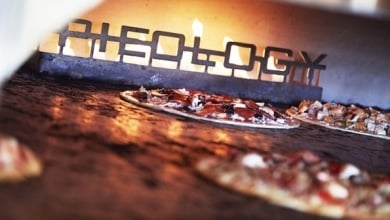 Pizzas in an oven with a three-dimensional metal logo inside the oven that spells out Pieology.  