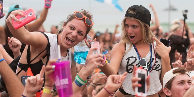 Two girls at an outdoor concert sitting on people's shoulders taking a selfie.