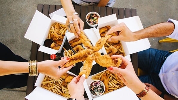 overhead view of four customers each a holding up a chicken finger, with meal boxes containing chicken fingers, fries and coleslaw on their table below. 