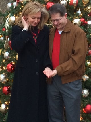 Sherri Johnston & David Wensil looking at engagement ring while standing in front of a Christmas tree. 