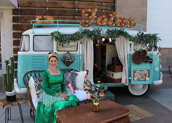 Queen Green wearing a green dress and a gold crown, sitting in front of an old VW van. 