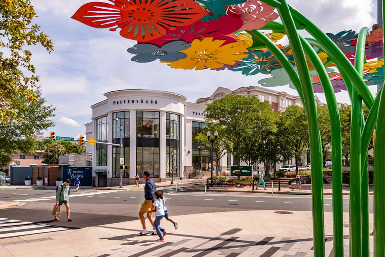 Intersection at The Crossing Clarendon with people crossing the street. Sculptural art is in the foreground and shops in the background.