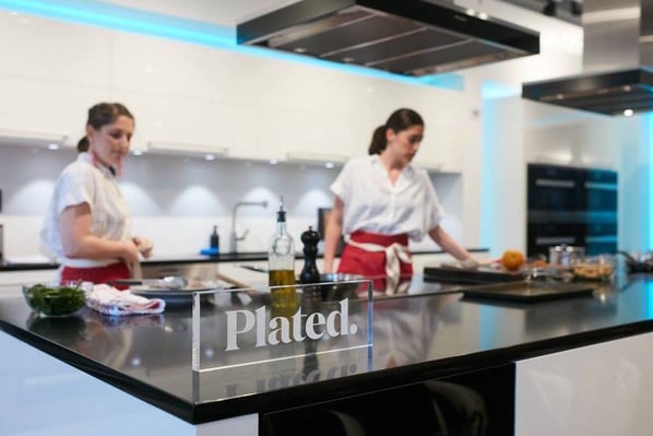 Two chefs in a kitchen with a glass Plated logo on a counter.