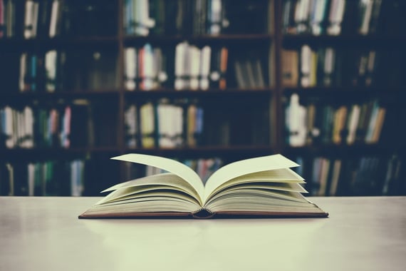 An open book on a table with shelves of books in the background. 