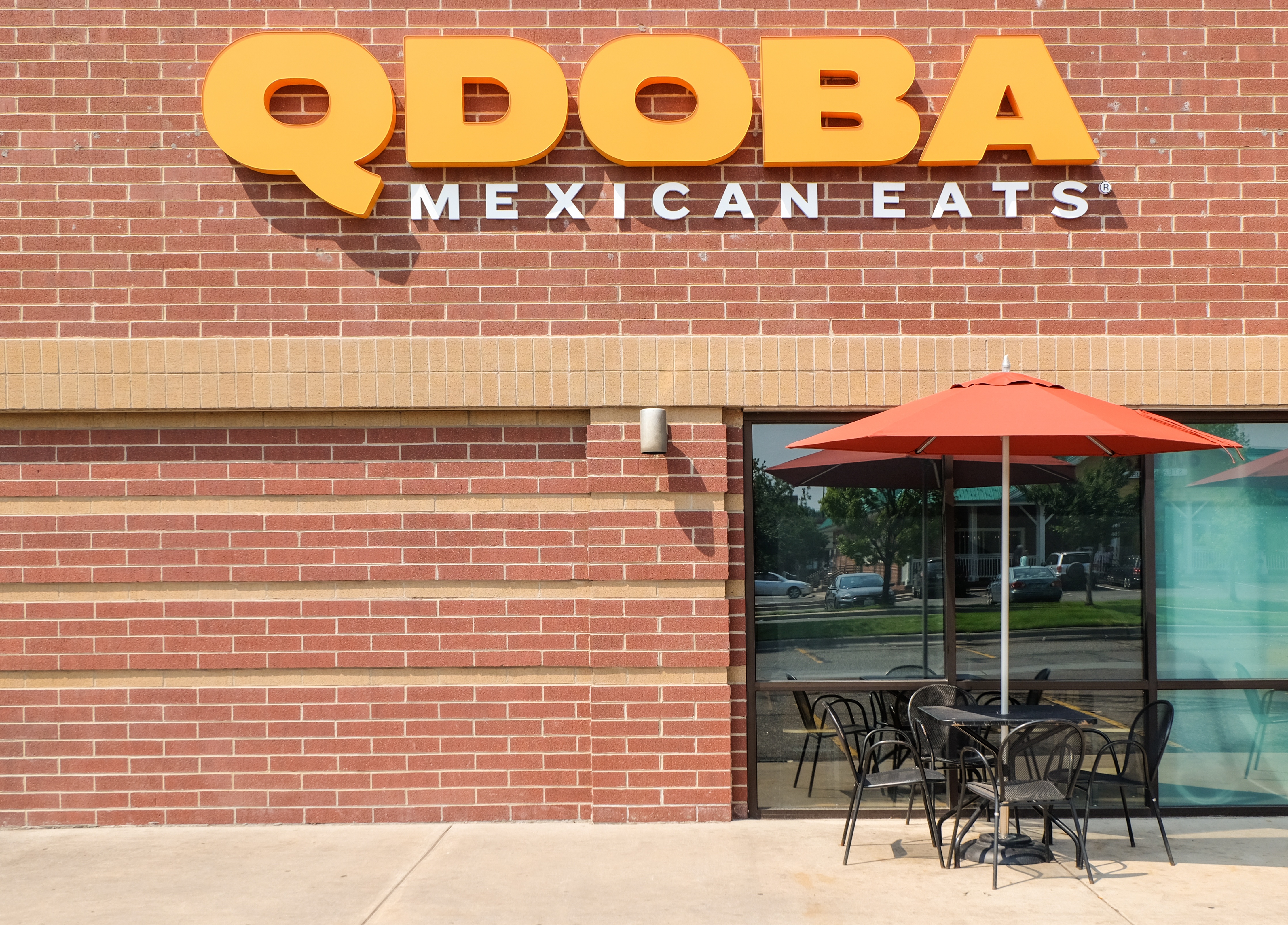 Qdoba signage on a brick wall with a table and chairs in front of it. 